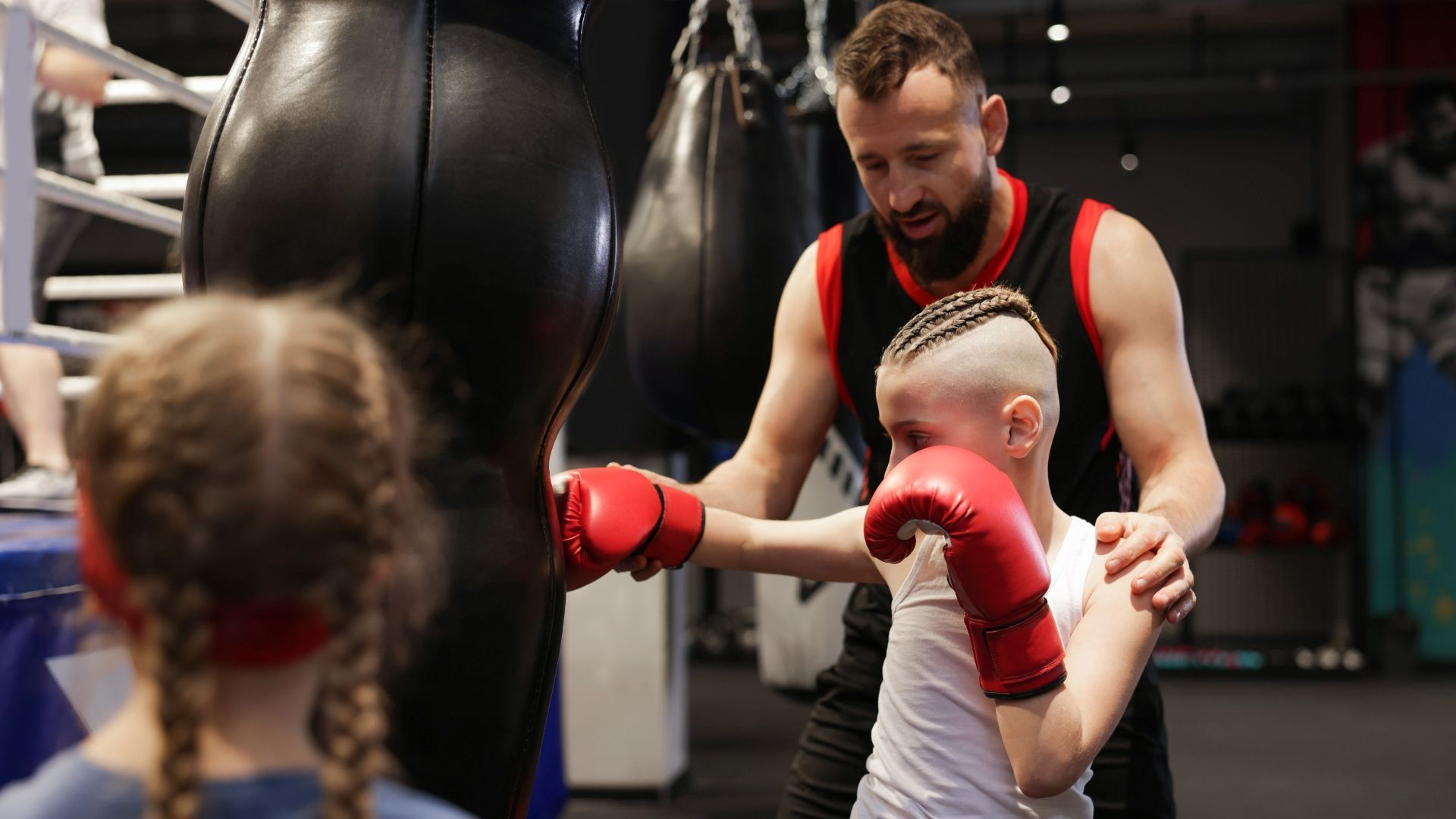 A coach assists a young person in boxing training, guiding their punch at a hanging heavy bag in a gym.