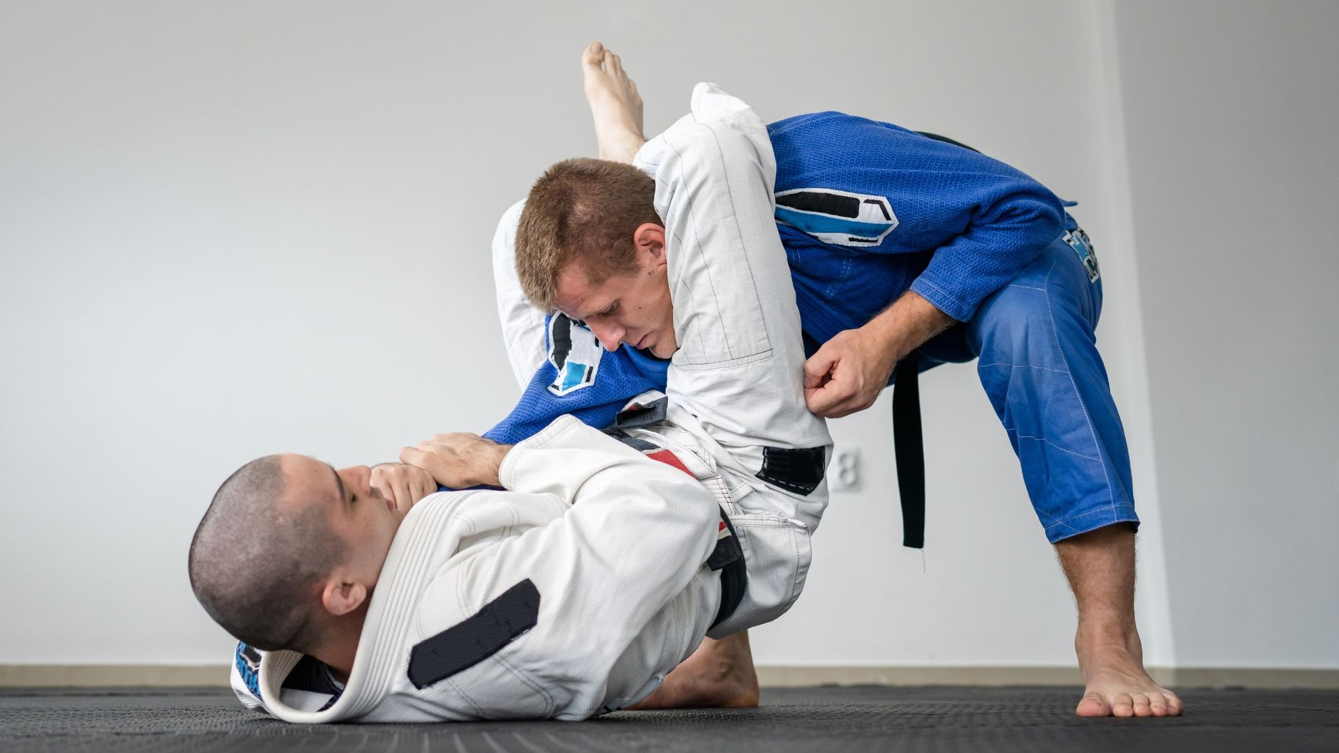 Two practitioners in martial arts gis perform a triangle choke maneuver on a mat in a bright, minimalist room.