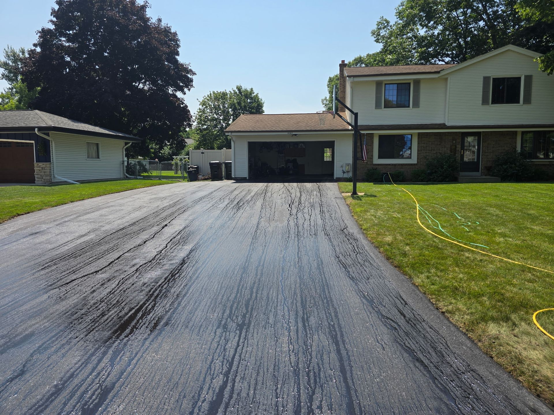 A freshly sealed black driveway leading to a two-story home with a garage on a sunny day.