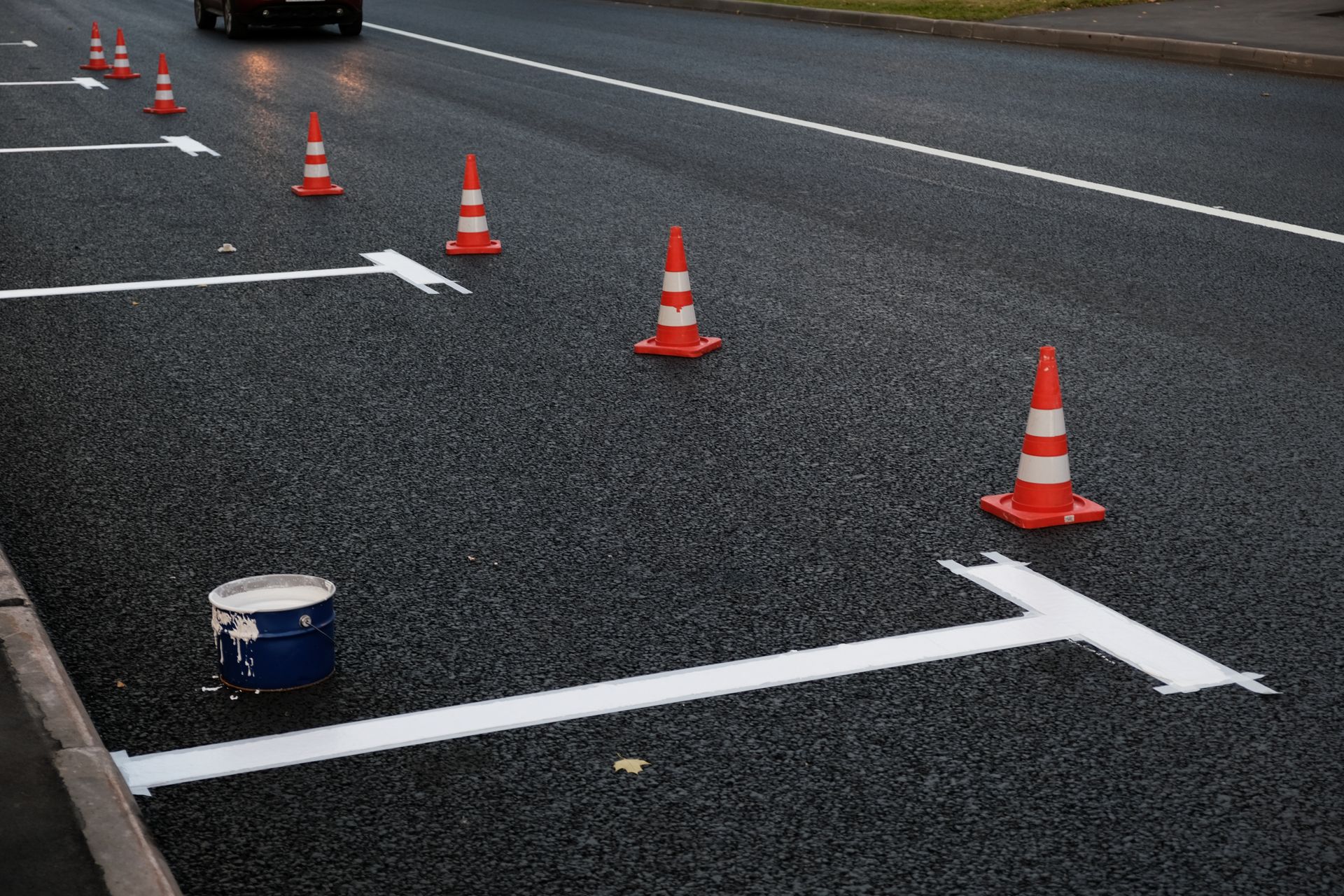Road markings being painted with white lines and orange traffic cones; a paint bucket sits nearby.