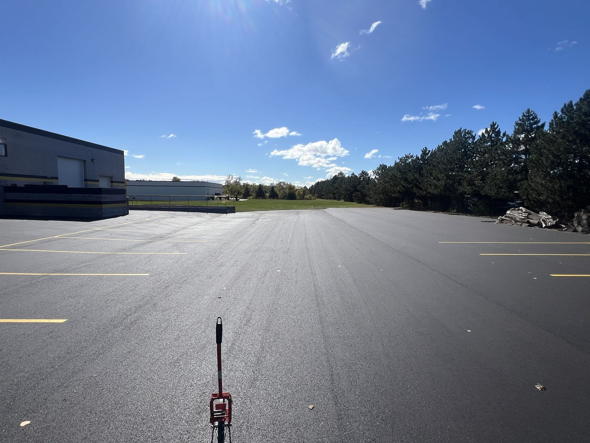 Empty sunlit parking lot beside a building and trees under a blue sky