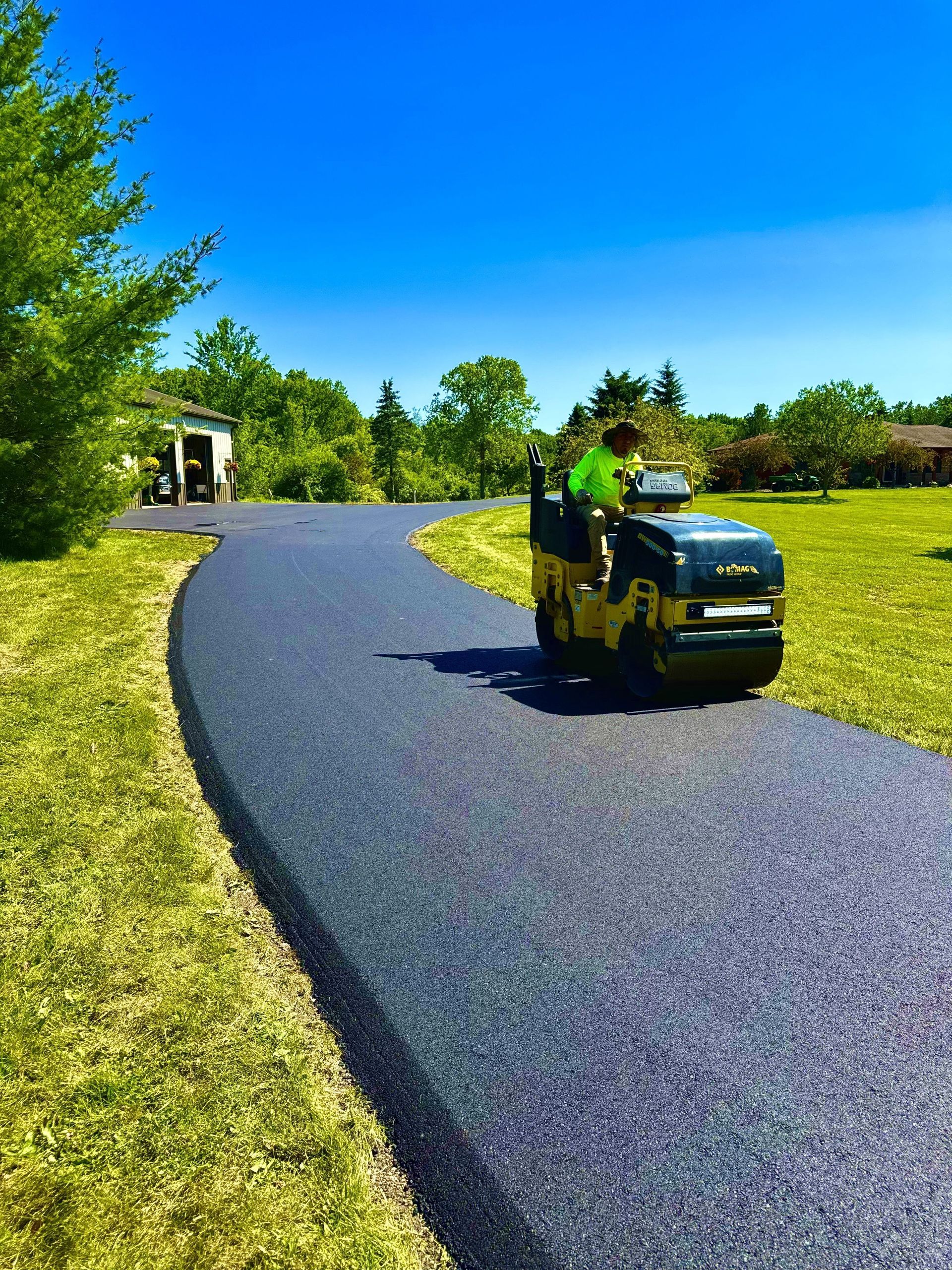 Black asphalt driveway being rolled by a yellow compactor on a sunny day.