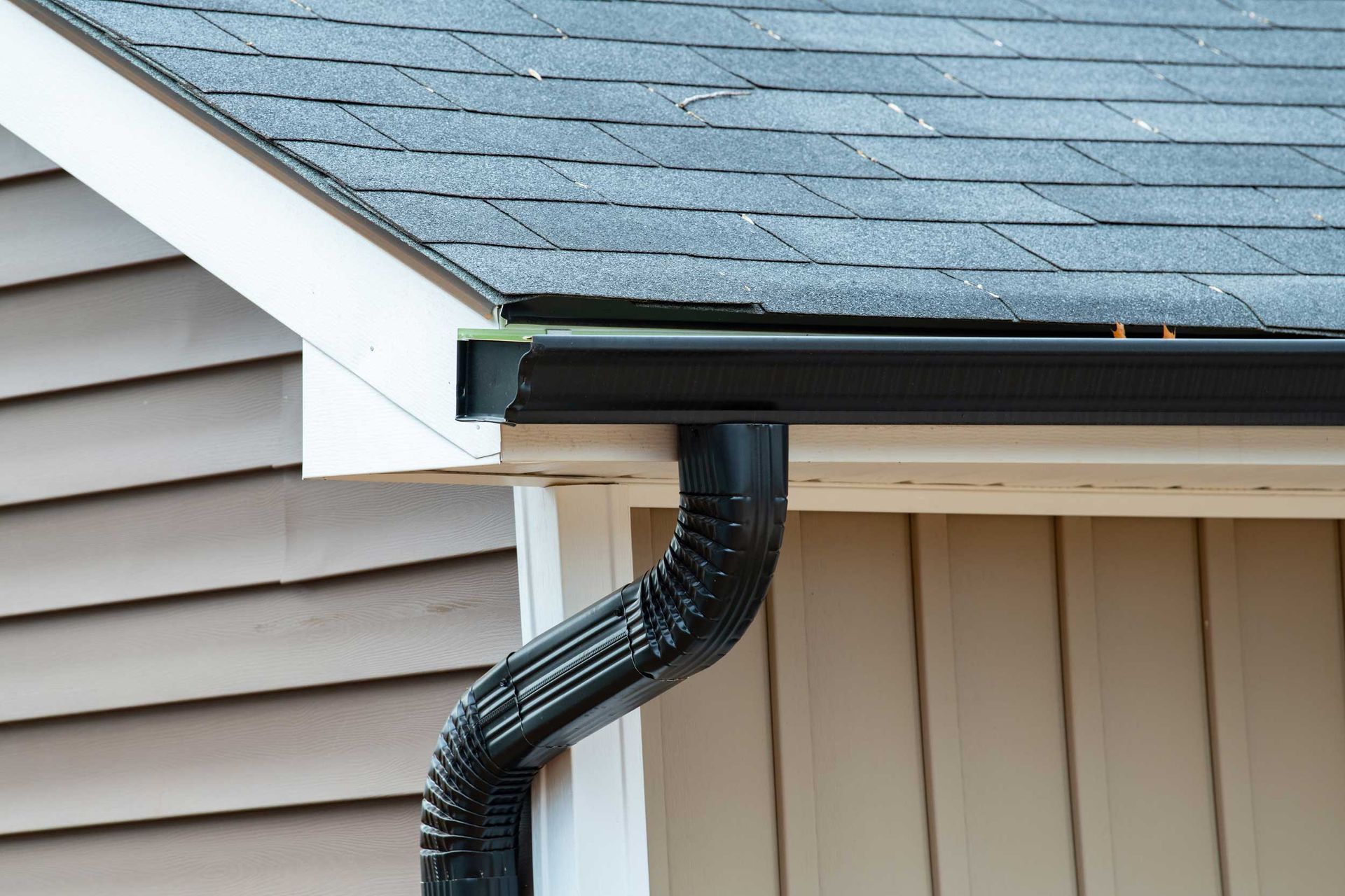 Black rain gutter attached to a roof with black shingles and a cream-colored exterior wall.