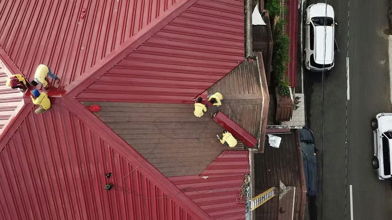 Workers on a red roof near a road with parked cars.