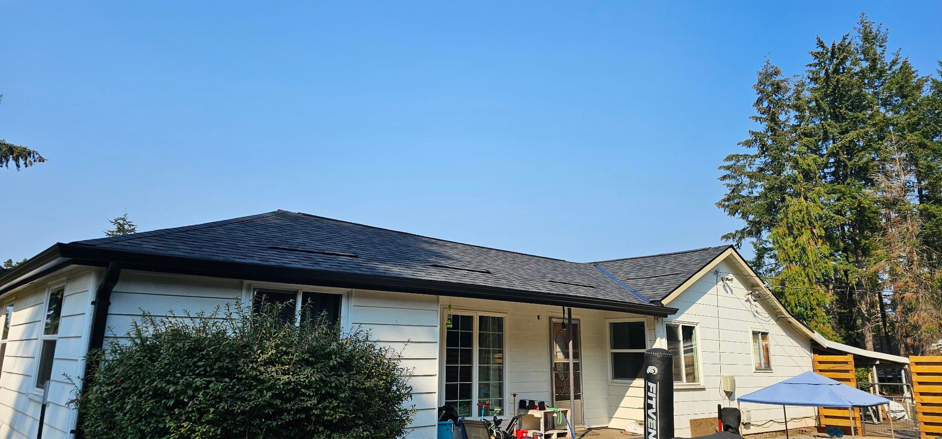 A white house with a dark roof under a clear blue sky. A green bush is in front of the house.
