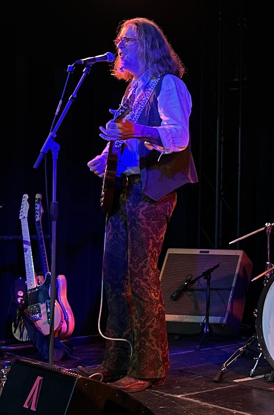French treedt op het podium op, gekleed in een vest en een broek met print. Hij heeft een microfoon en banduitrusting bij zich.