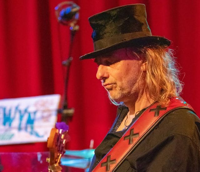 The Fish met zwarte hoed speelt basgitaar op podium tegen rode achtergrond.