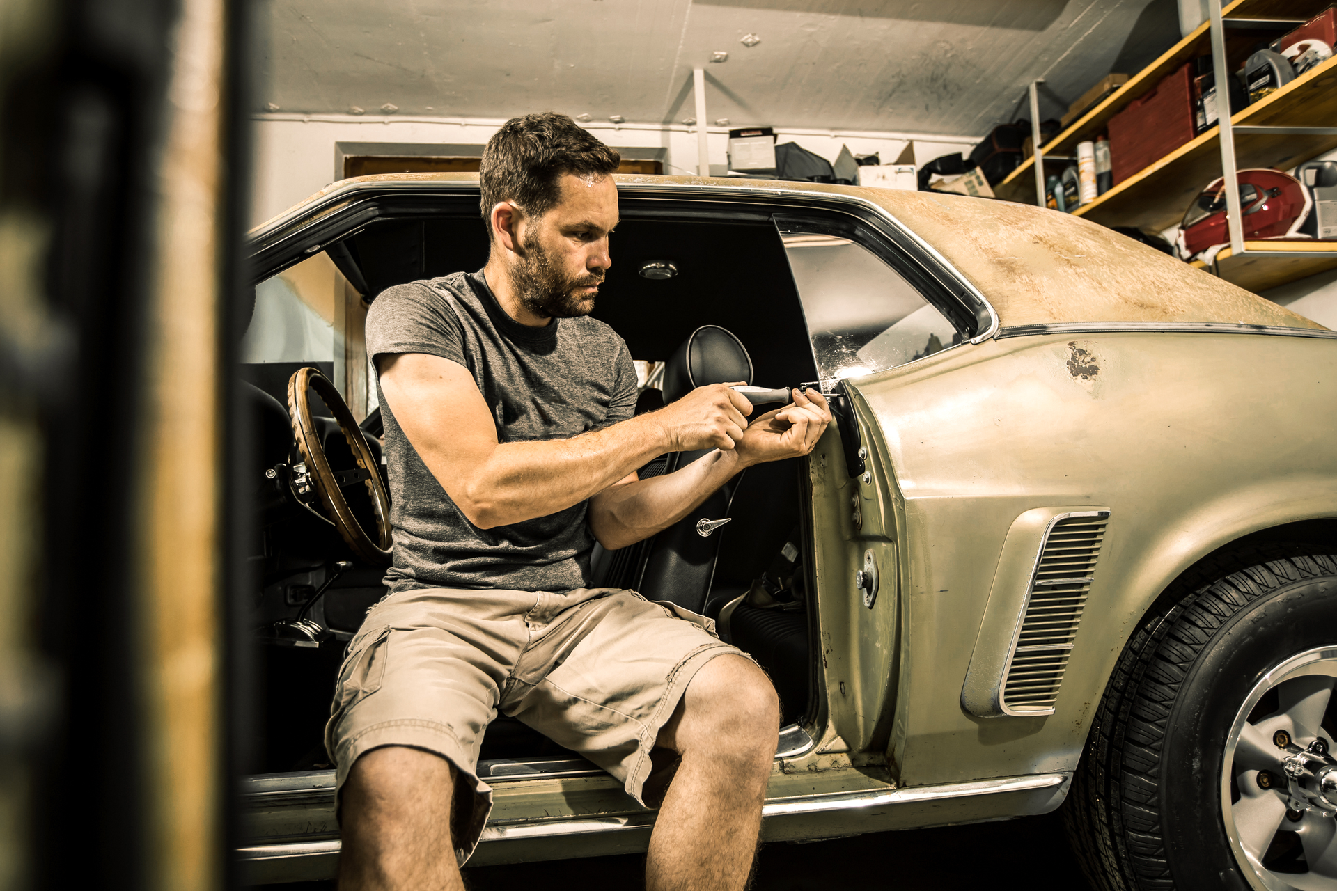 A man is sitting in a car in a garage working on it.