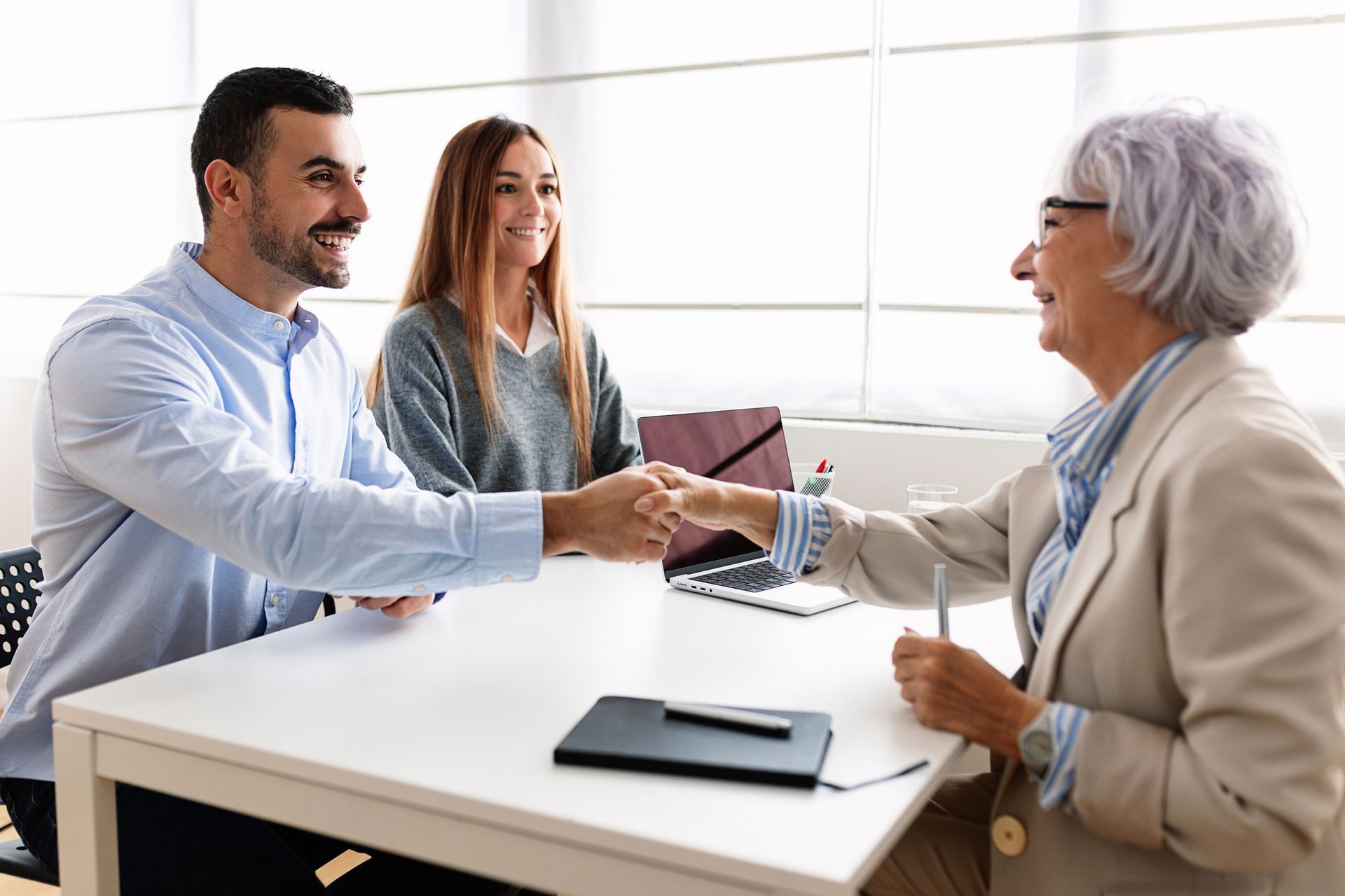 Two people meeting with an advisor, shaking hands across a desk with a laptop and notebook.