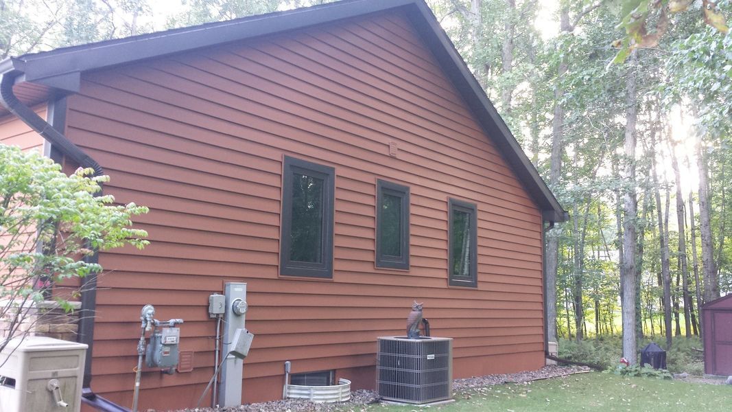 Wood-sided cabin exterior in a wooded yard, with utility meters and windows along the wall.