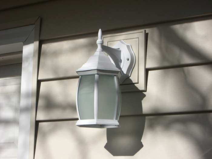 White outdoor wall lantern on beige house siding, casting a shadow in sunlight