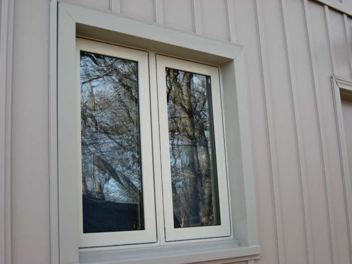 White-framed double window on a light-sided building, reflecting trees outside.