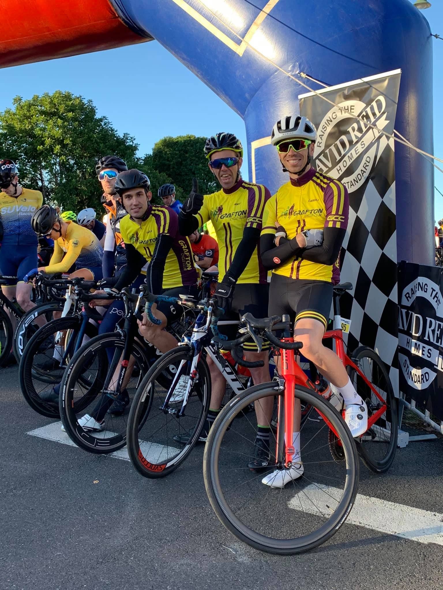 Cyclists in yellow jerseys pose with their bikes at a race start line under an arch — Grafton City Cycles in Grafton, NSW