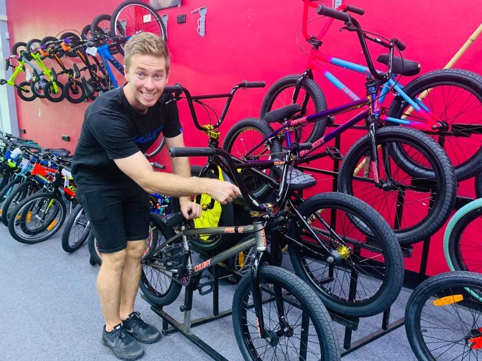 Man smiles, points at black BMX bike in bike shop. Bikes on display; red wall background — Grafton City Cycles in Grafton, NSW