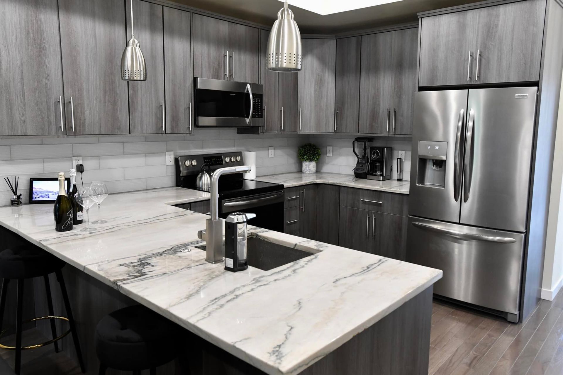 Modern kitchen with gray cabinets, marble countertops, stainless steel appliances, and silver pendant lights.