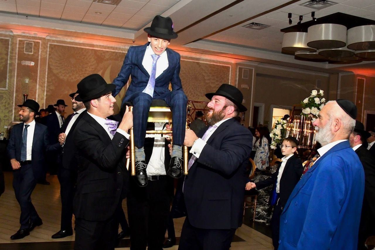 People in suits and hats lift a seated person on a chair in a brightly lit event hall.