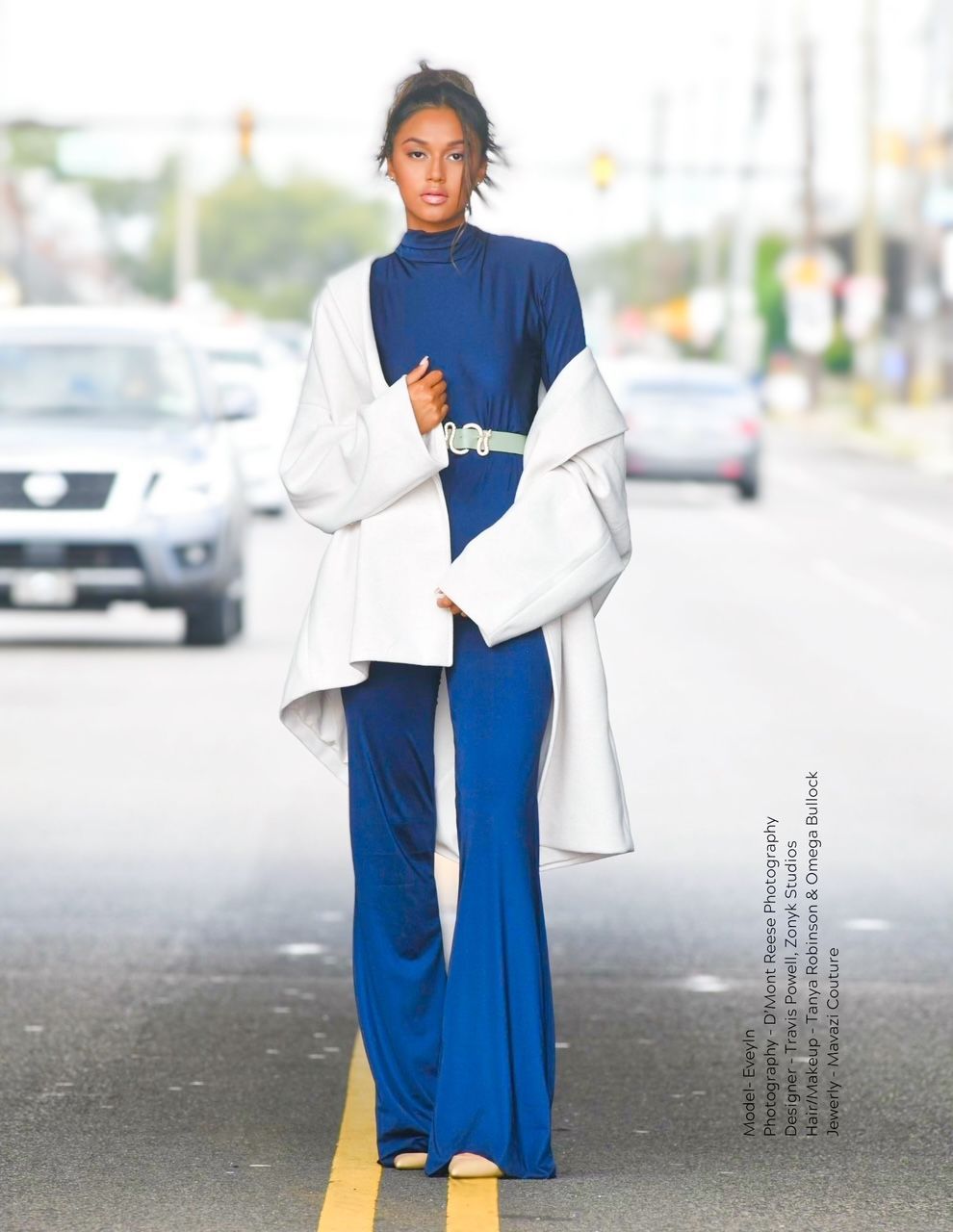 Woman in blue jumpsuit and white coat stands in the middle of a street, cars blurred in the background.