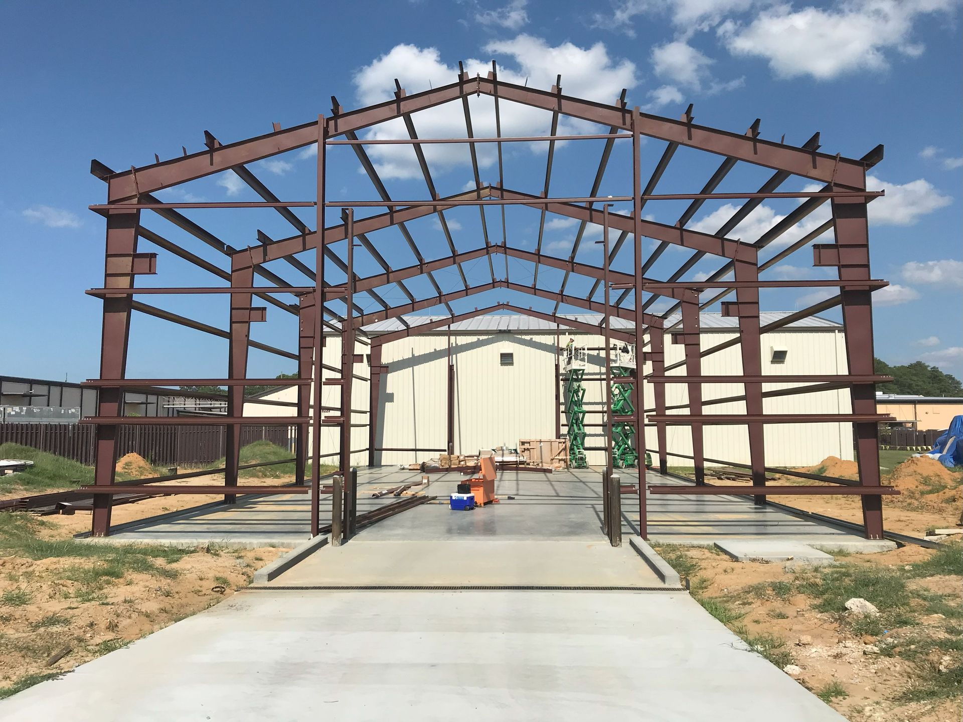 Steel frame of a building under construction with brown beams, sitting on concrete, with tan building in the background.