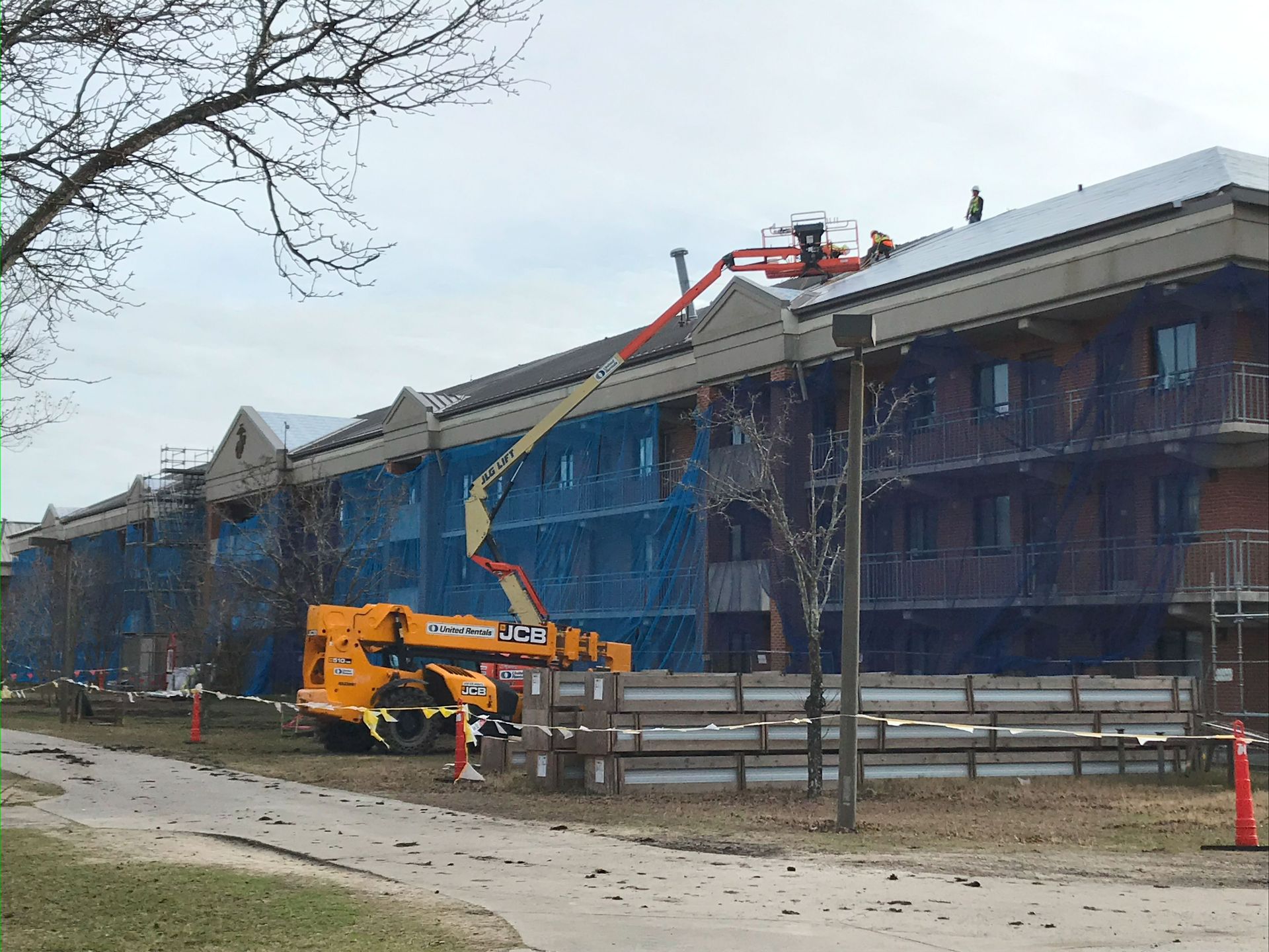 Building under construction; scaffolding, lift, workers on the roof; blue netting; overcast day.