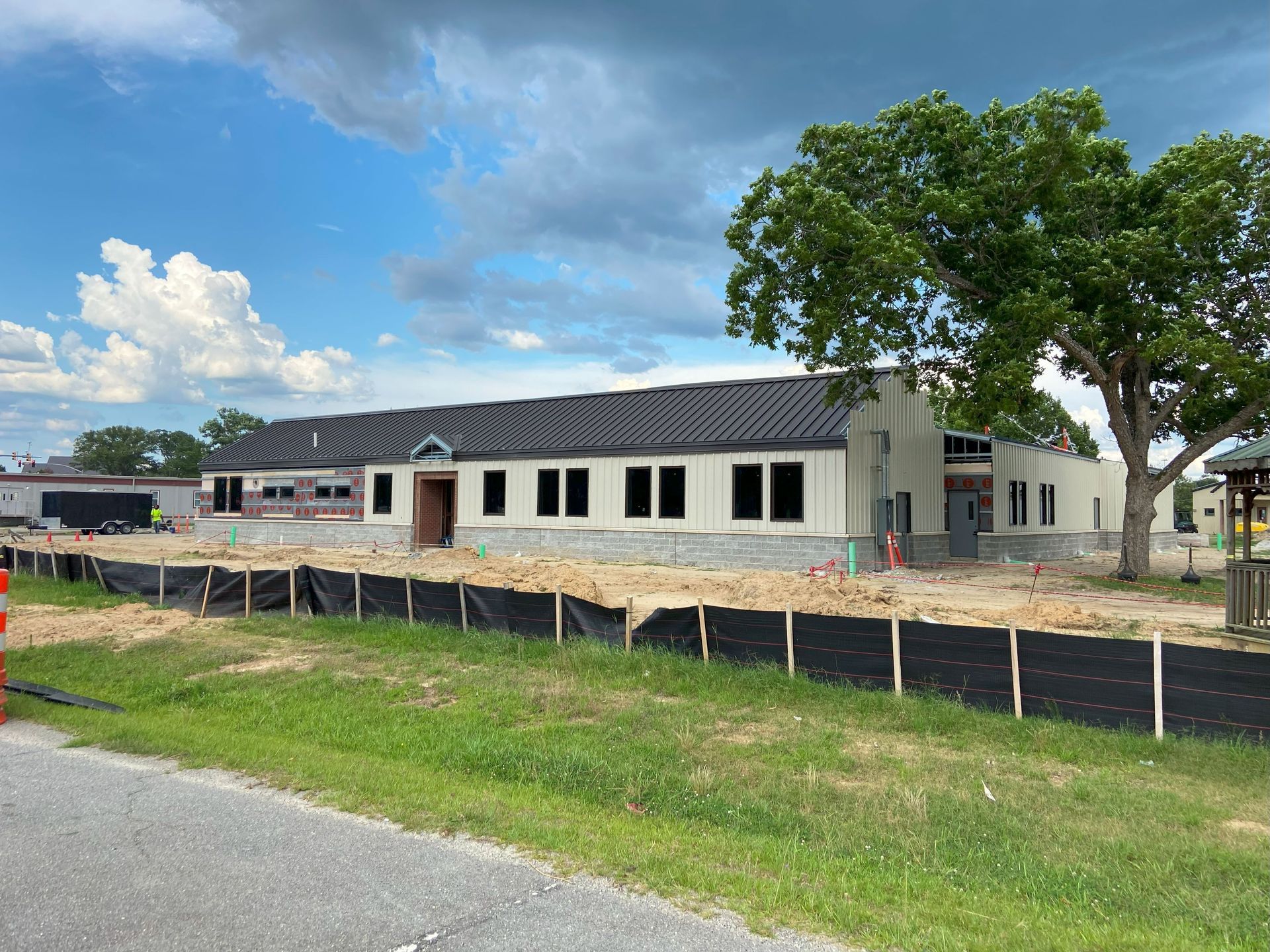 Building under construction with black roof, light walls, and tree, surrounded by a black barrier and green grass.