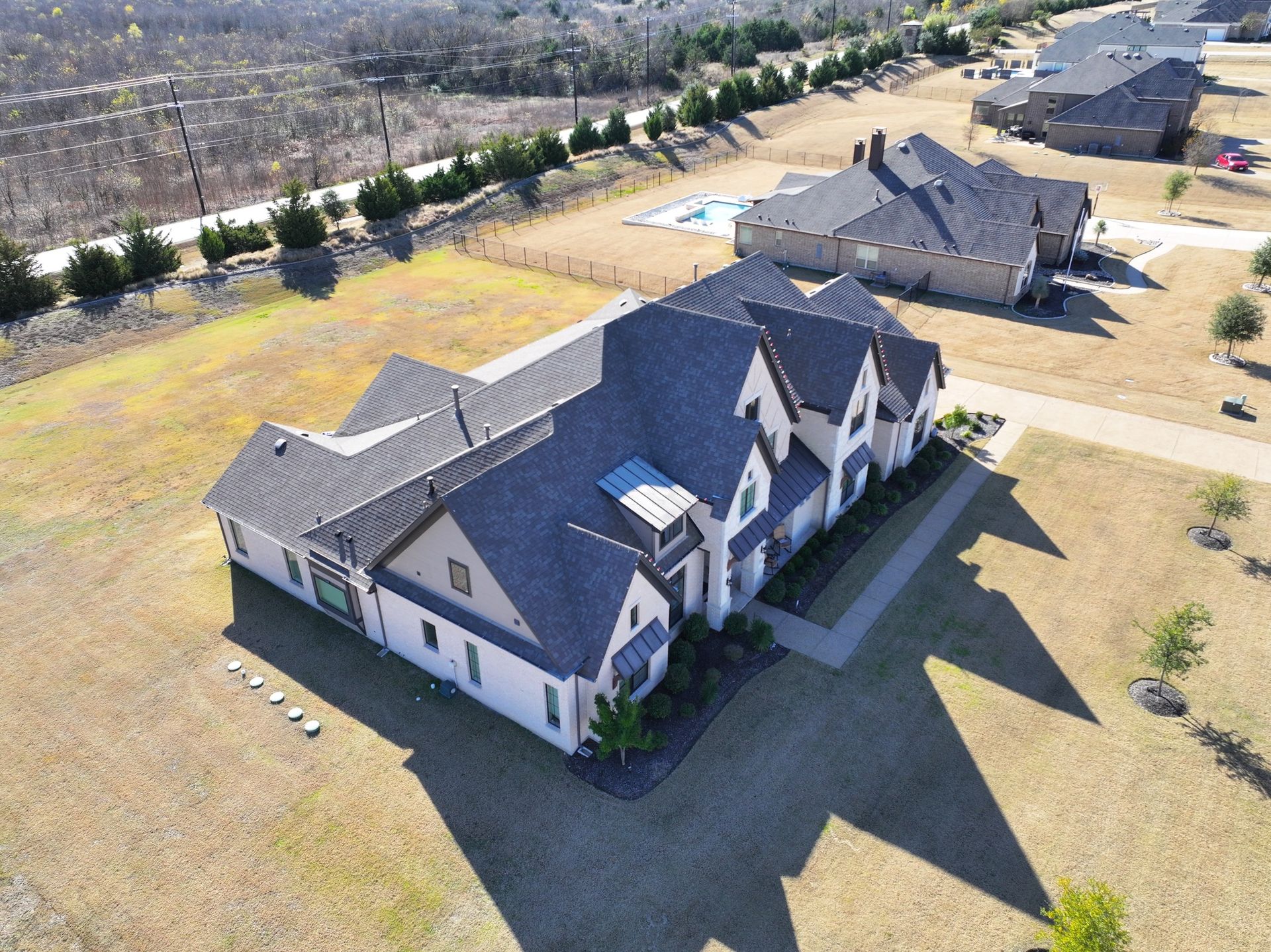 Aerial view of a large house with a complex roof, casting a long shadow on dry grass.