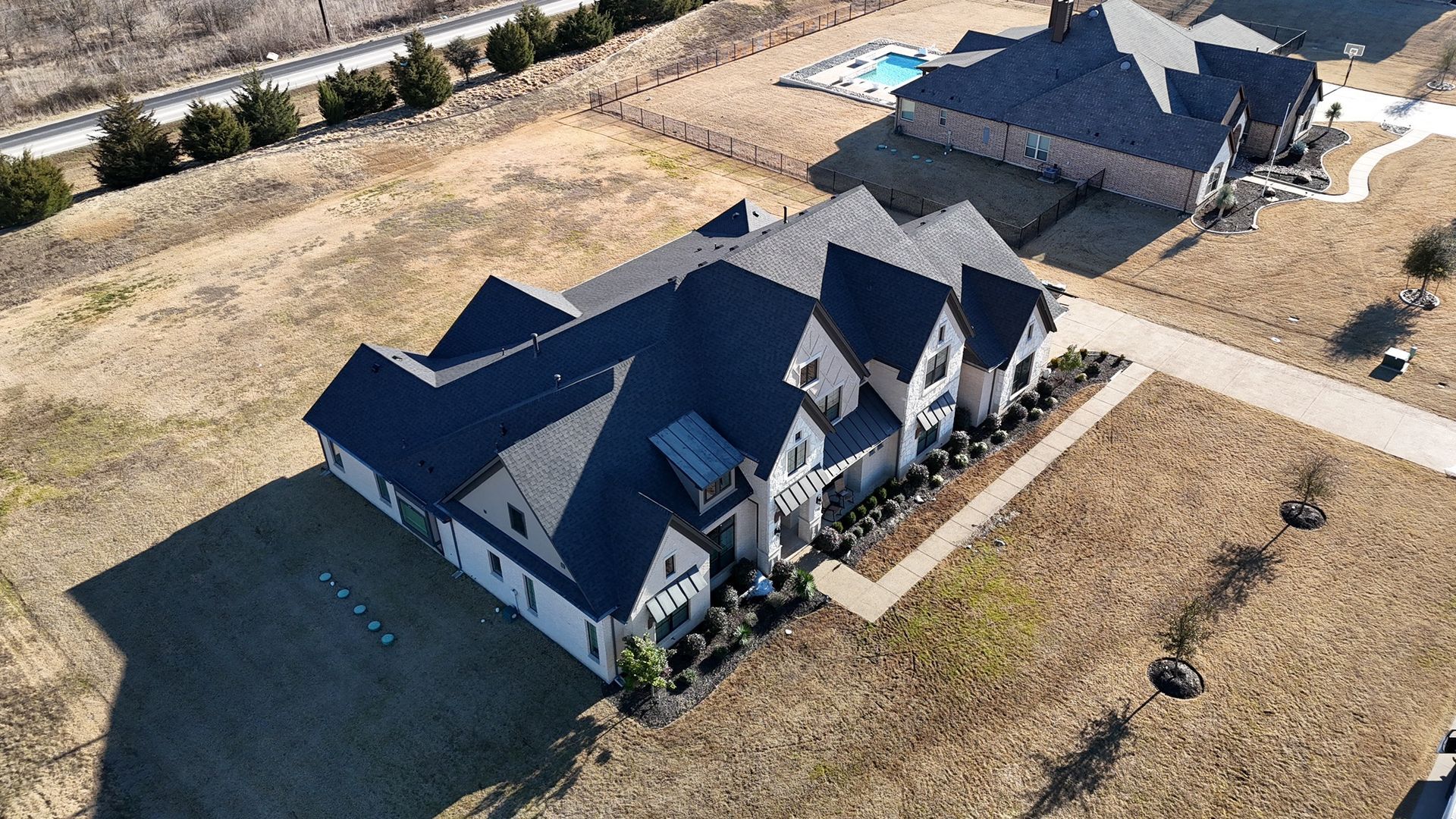 Aerial view of a large, dark-roofed house with a long driveway and surrounding dry grass.