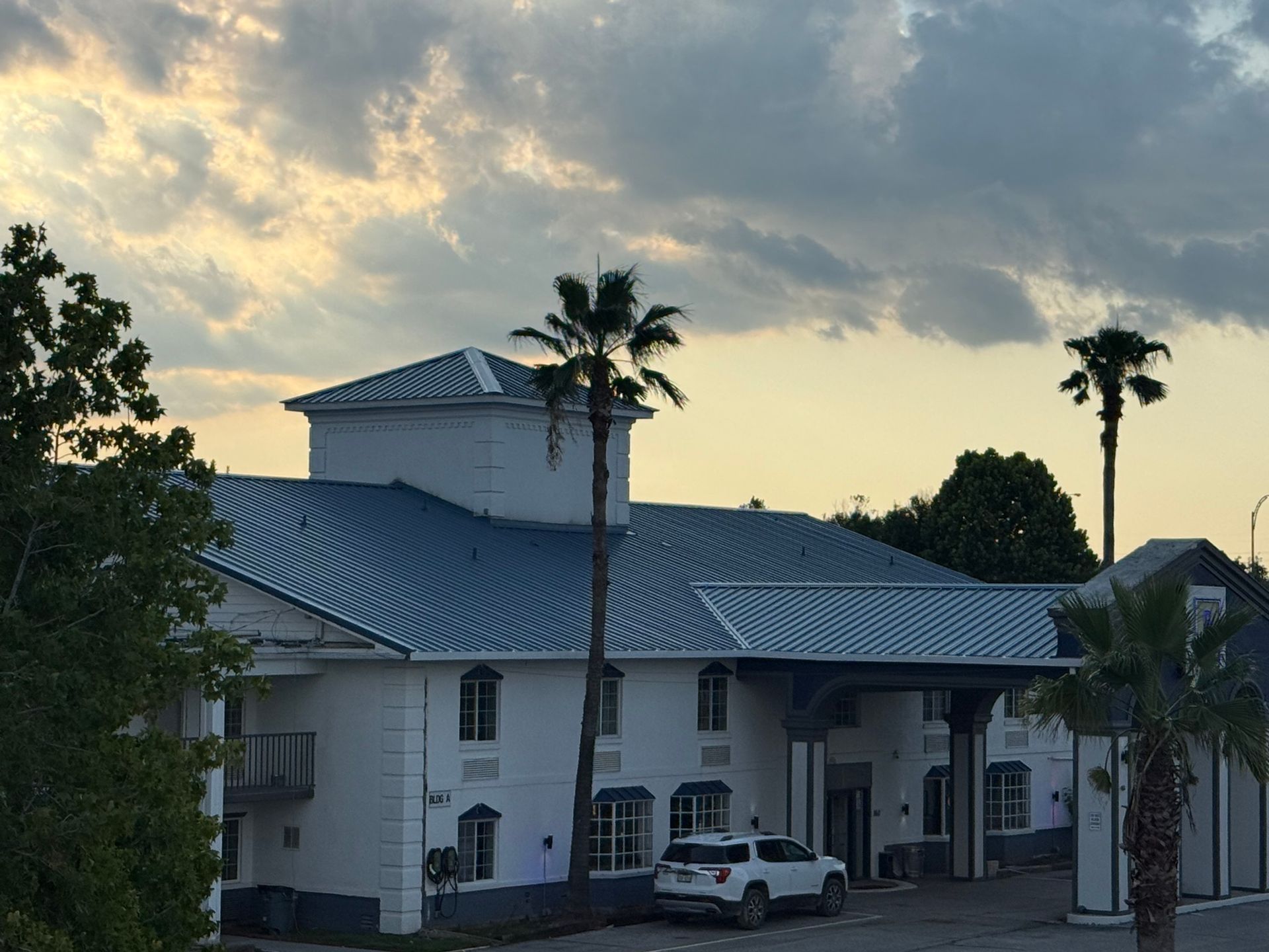 White motel with a blue roof, palm trees, and a car parked out front at dusk.