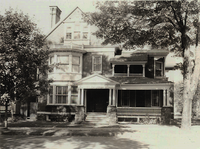 Black and white photo of a large, two-story house with a porch and trees in the front yard.