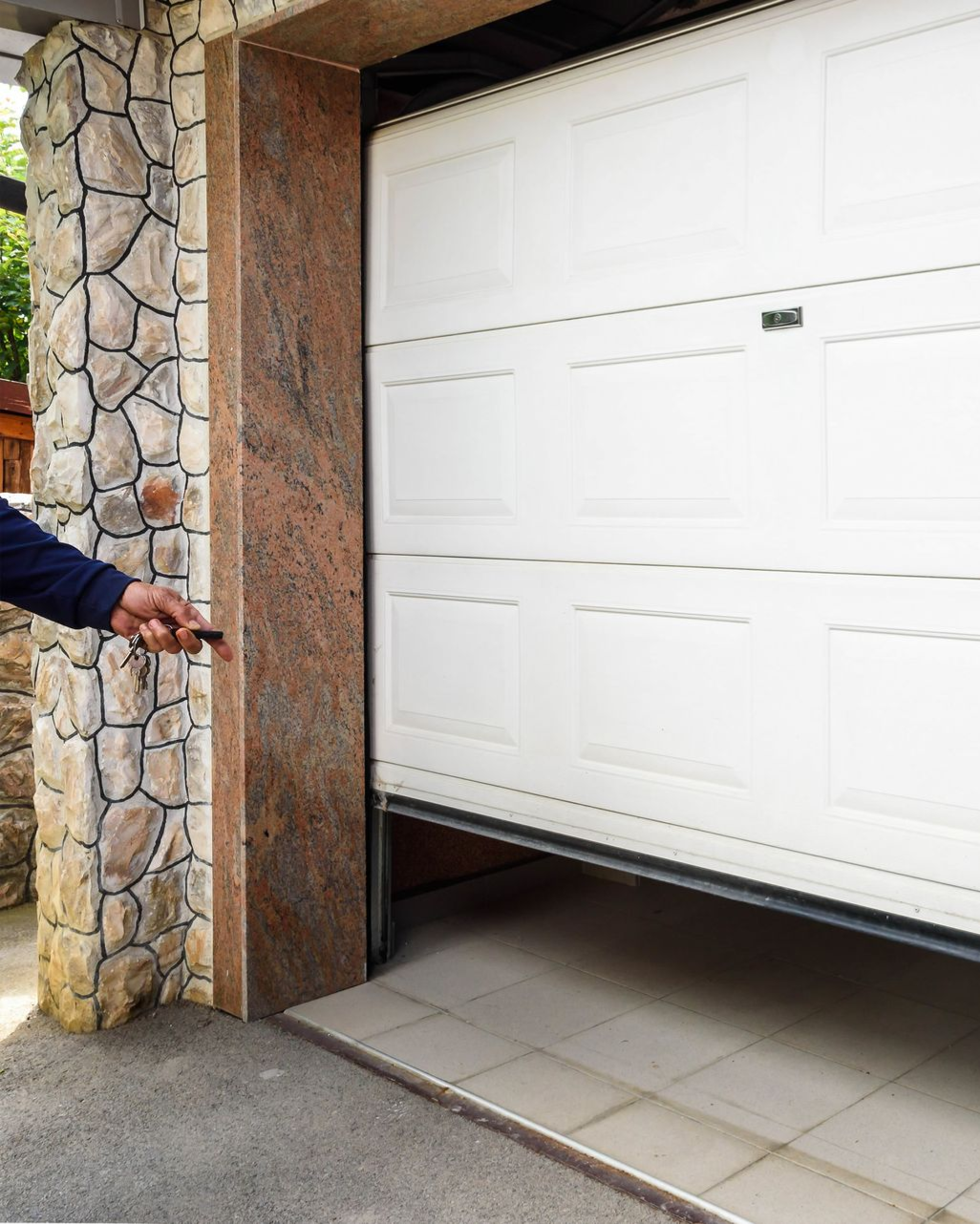 Garage door opening, controlled by a remote. Person in blue sleeve, stone and granite facade.
