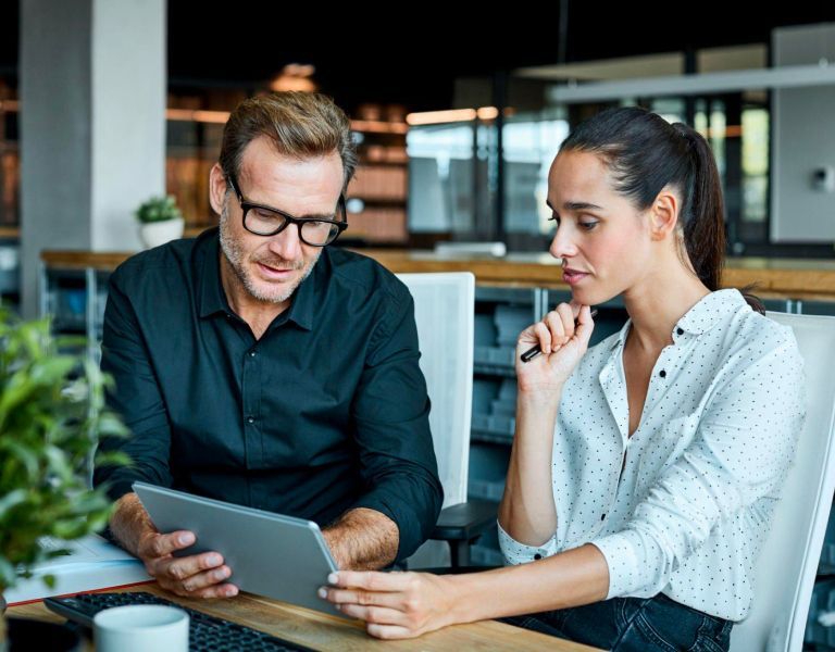 A man and a woman are sitting at a table looking at a tablet.