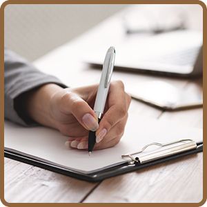 Woman's hand writing on a clipboard with a pen; on a desk with a laptop and paper.
