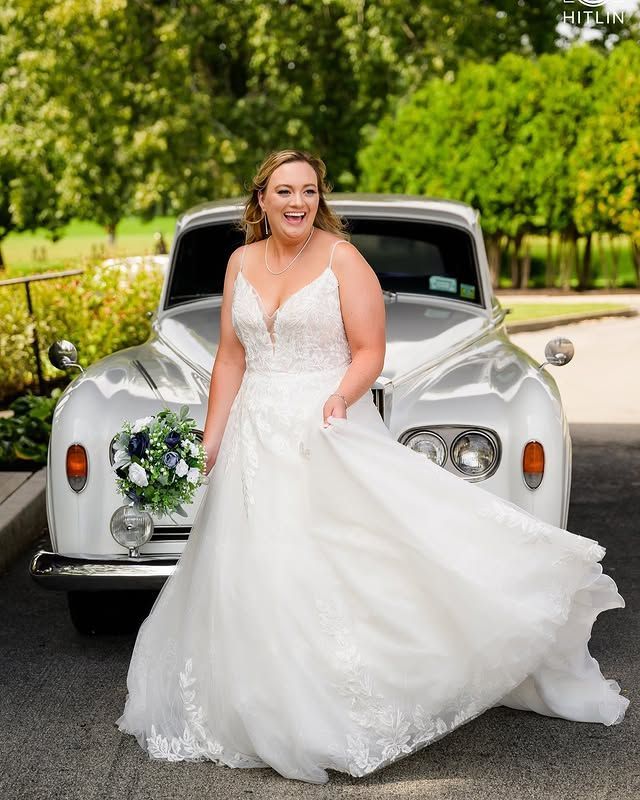 A woman in a wedding dress is standing in front of a white car.