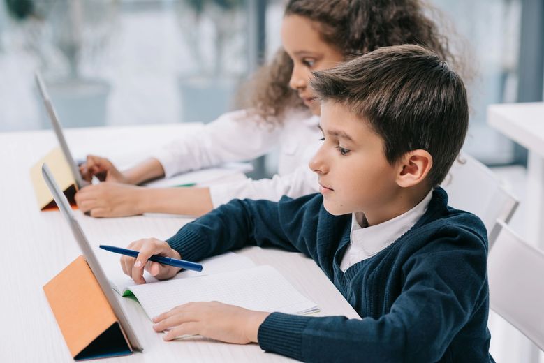 Un niño utiliza una tableta en un aula, mientras otro estudiante trabaja al fondo.