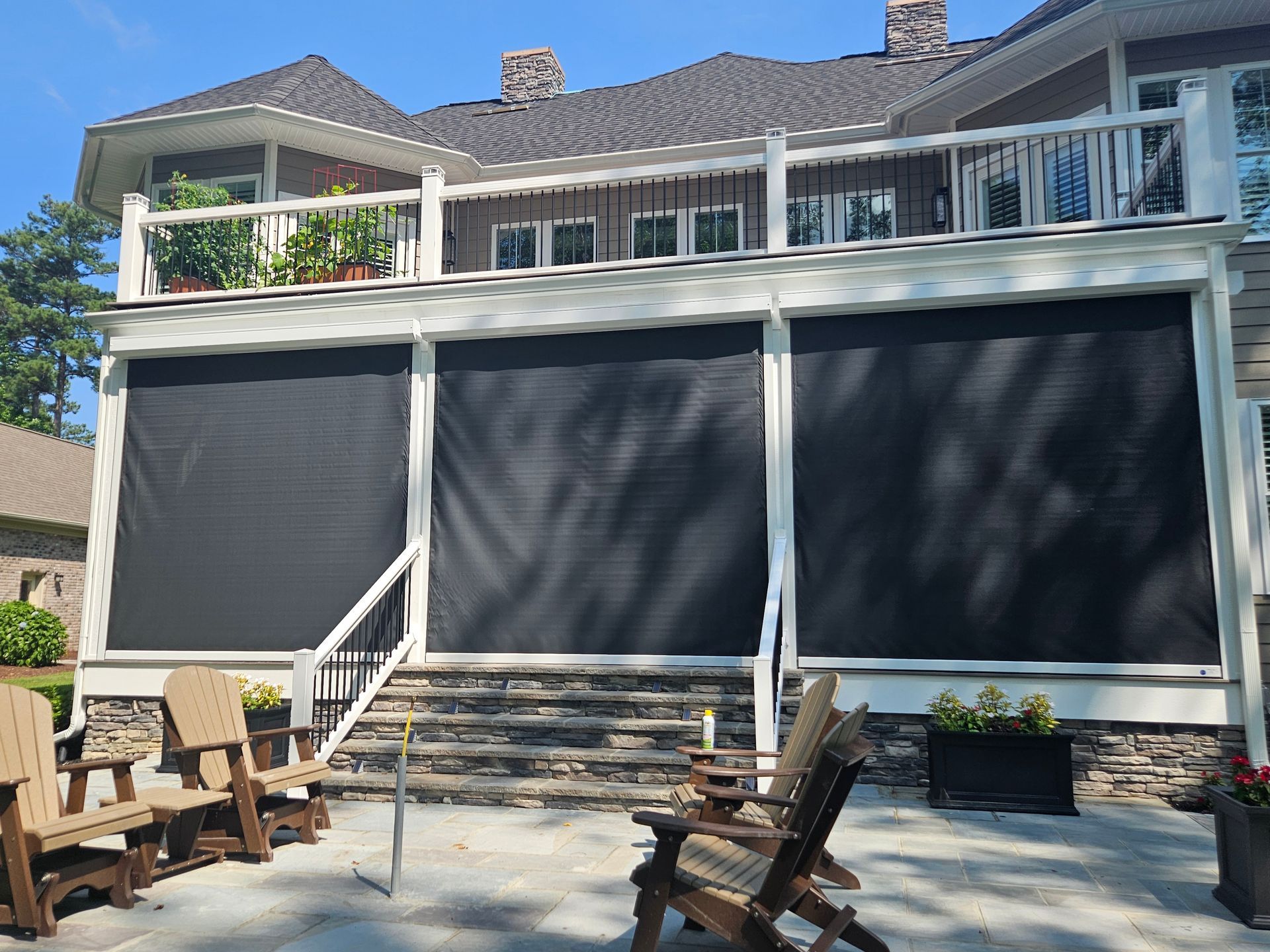 Outdoor patio with black sun shades, wood chairs, and a house in the background.