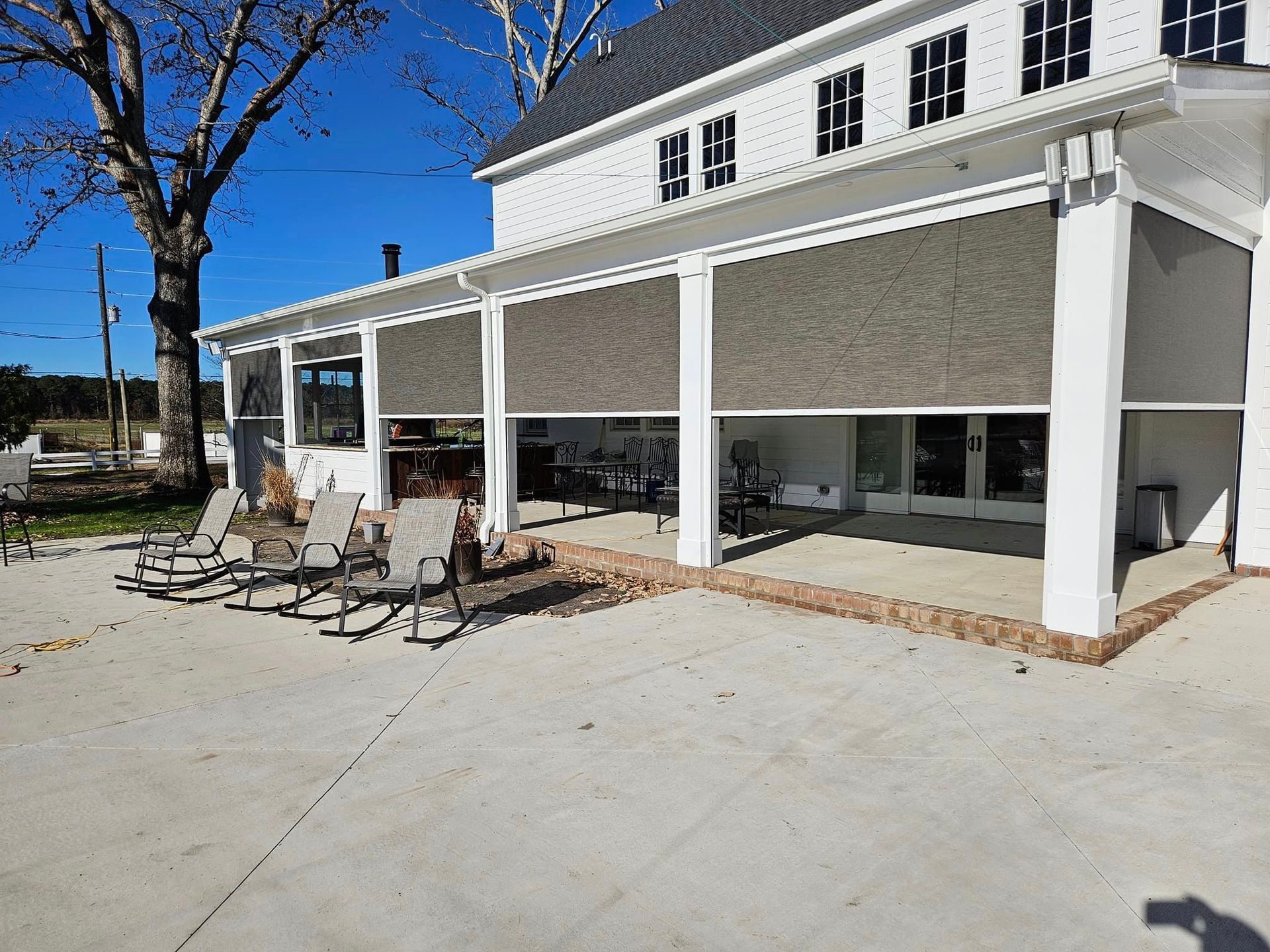 A large white house with a covered patio with chairs and a tree in the background.