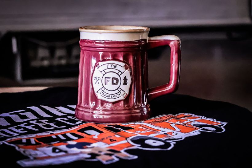 Burgundy ceramic mug with firefighter emblem, sitting on a black shirt with orange design.
