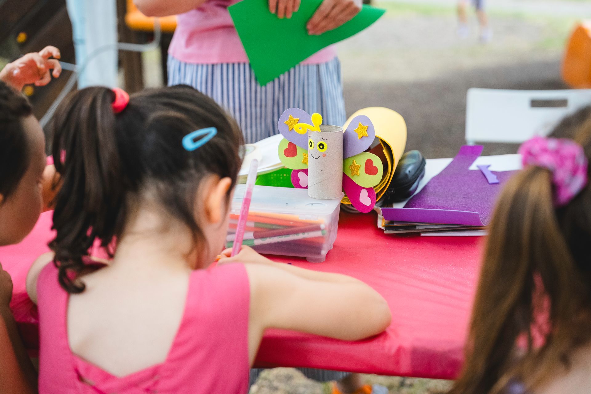 Bambini che lavorano a un tavolo rosa all'aperto. Sono visibili materiali per il bricolage e una donna con un foglio di carta verde.