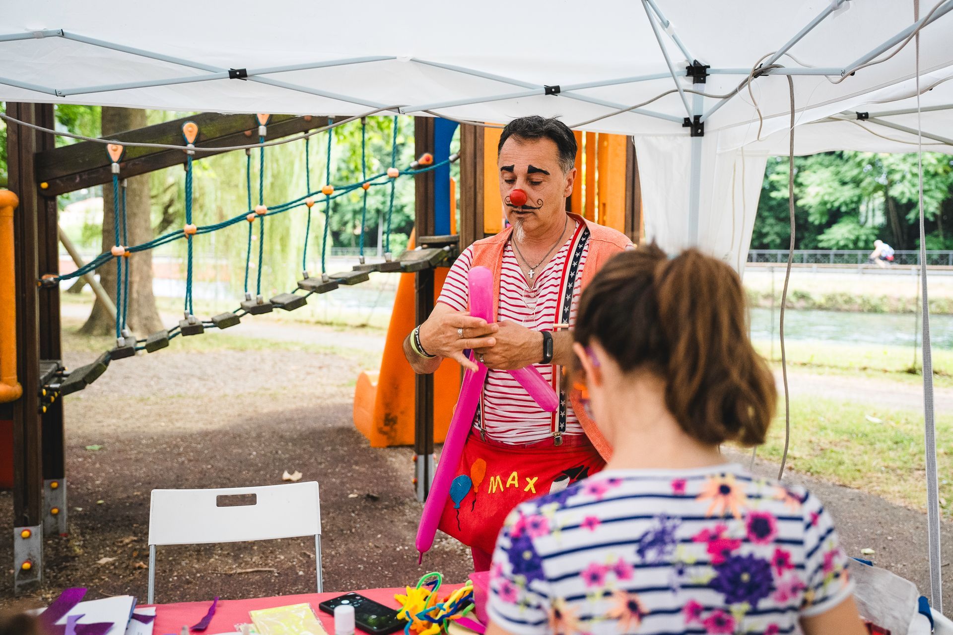 Un clown con il naso rosso e le bretelle crea un palloncino rosa a forma di animale per un bambino durante un evento all'aperto.
