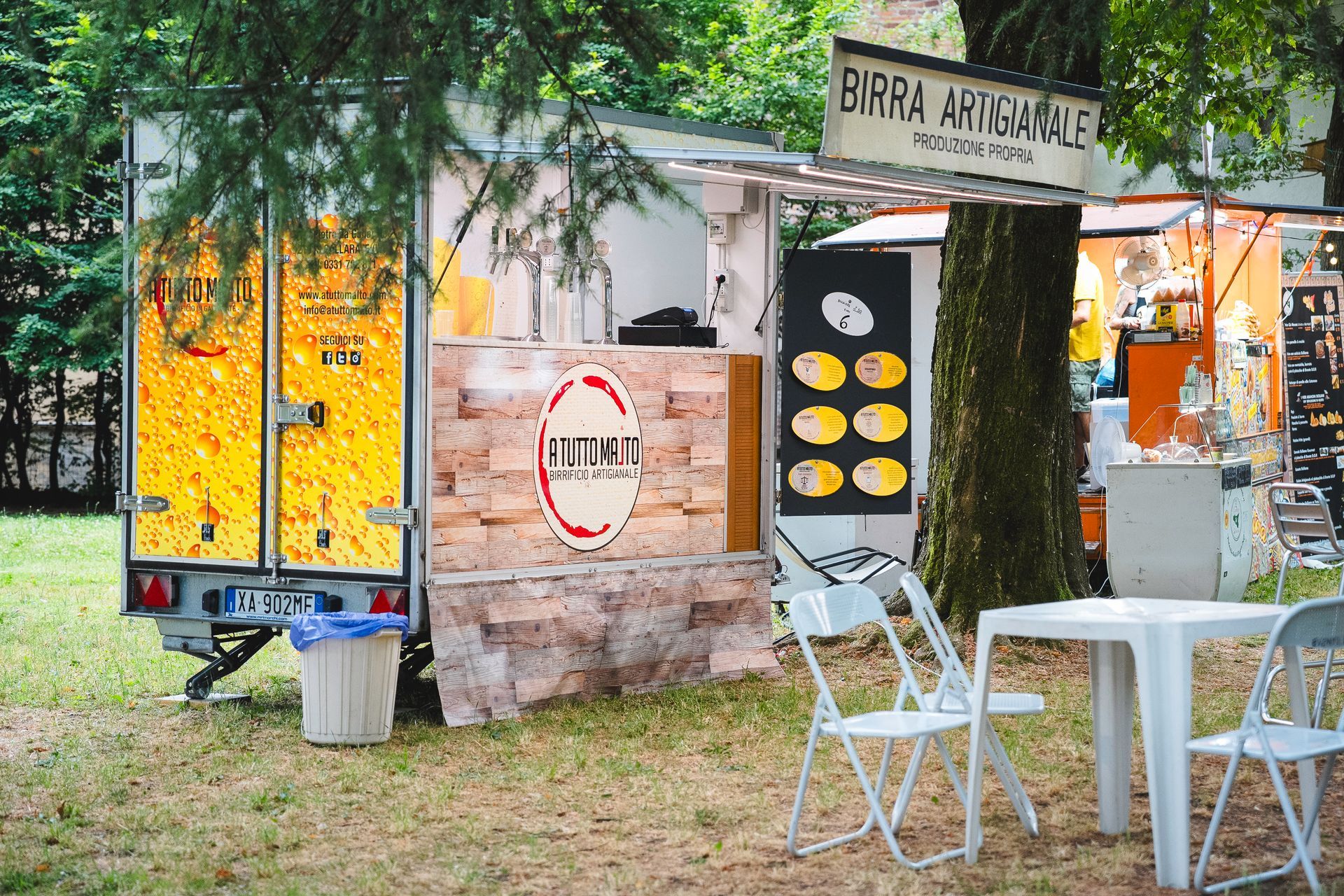 Camion di cibo con facciata in pietra, che serve birra e cibo; all'esterno sono allestiti tavoli e sedie bianchi.