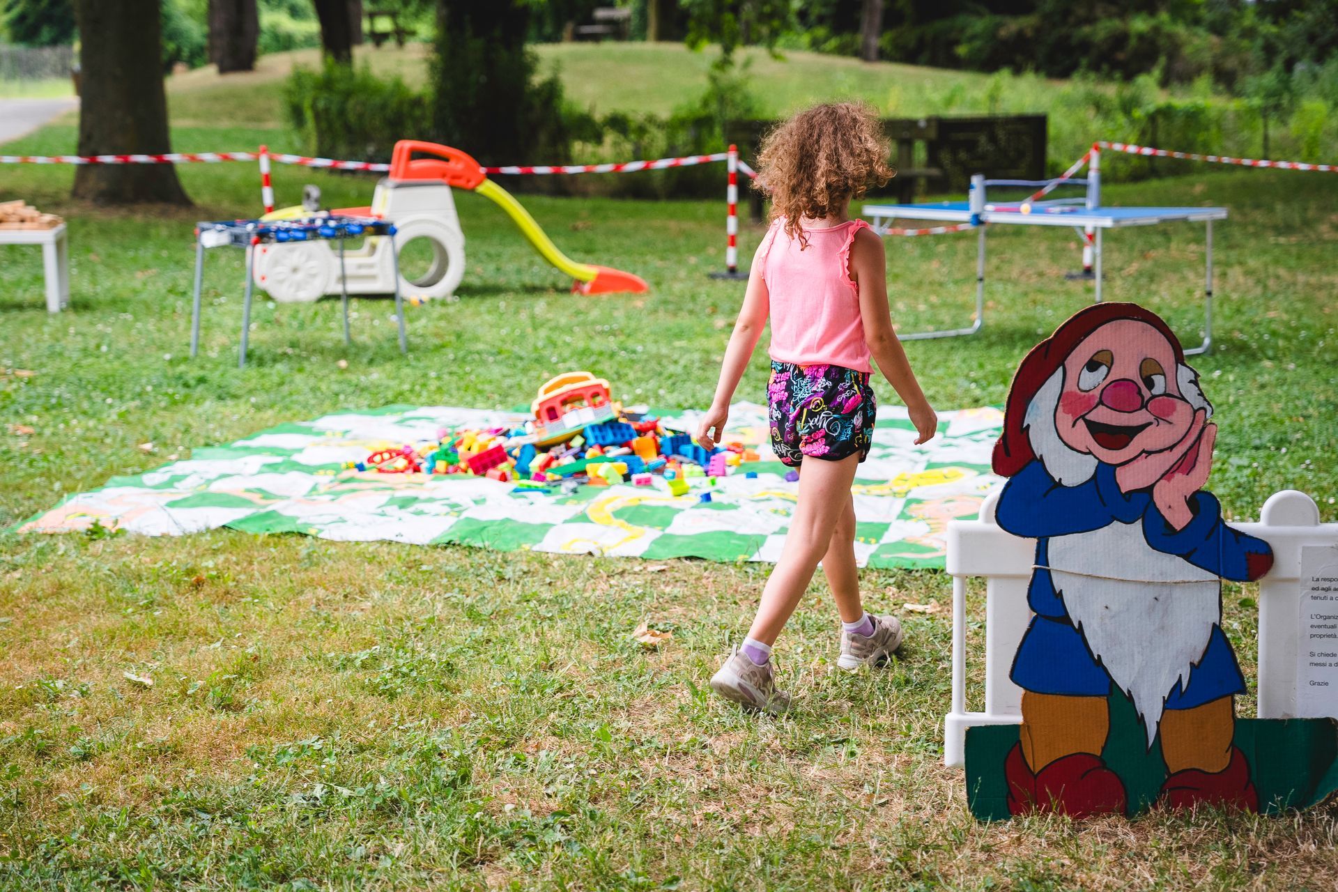 Una bambina cammina verso un'area giochi con uno scivolo e dei giocattoli, in un parco con la sagoma di un nano.