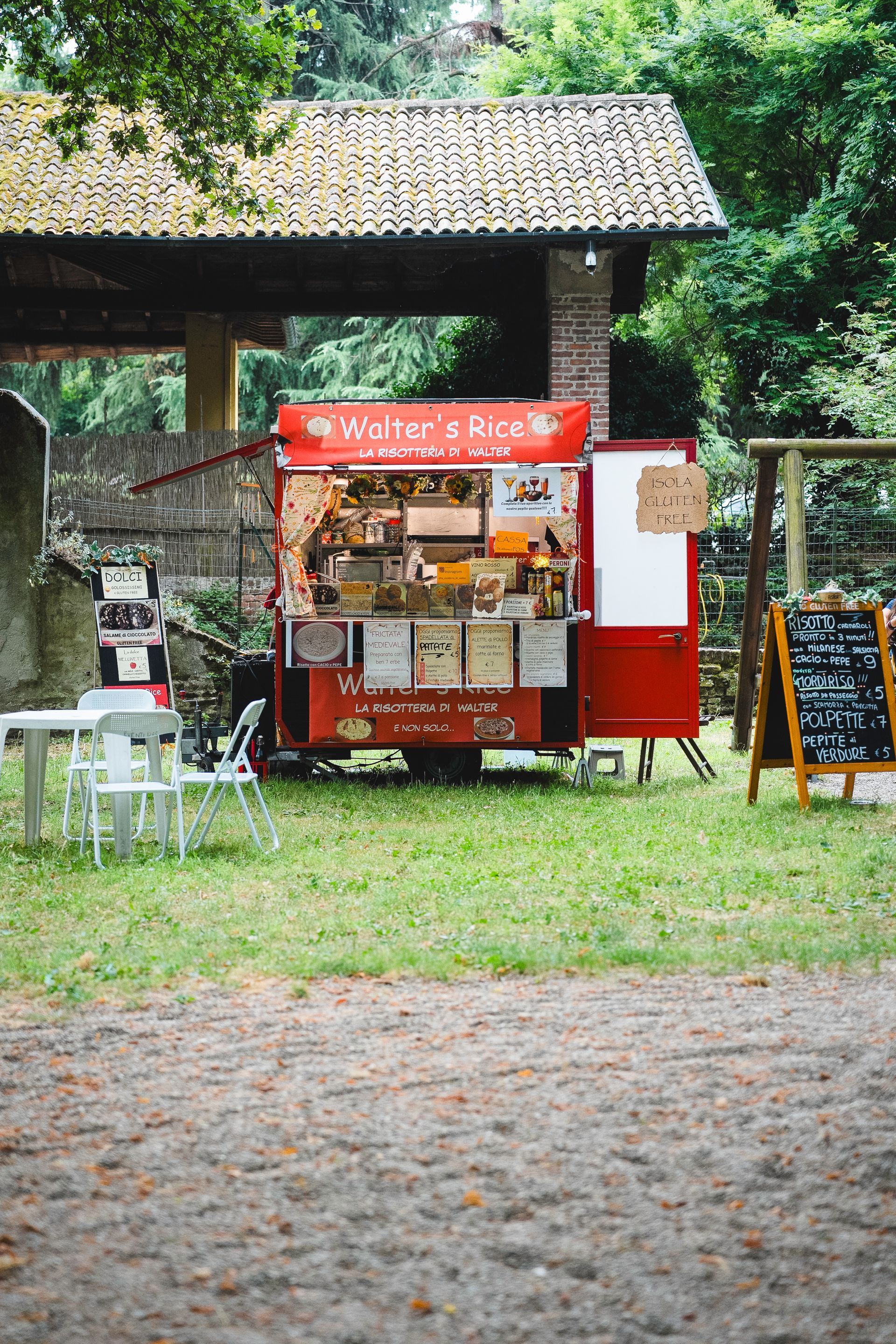 Camion rosso con cibo in un'area erbosa con tavoli. Espone il menu e vende cibo. Alberi ed edifici sullo sfondo.