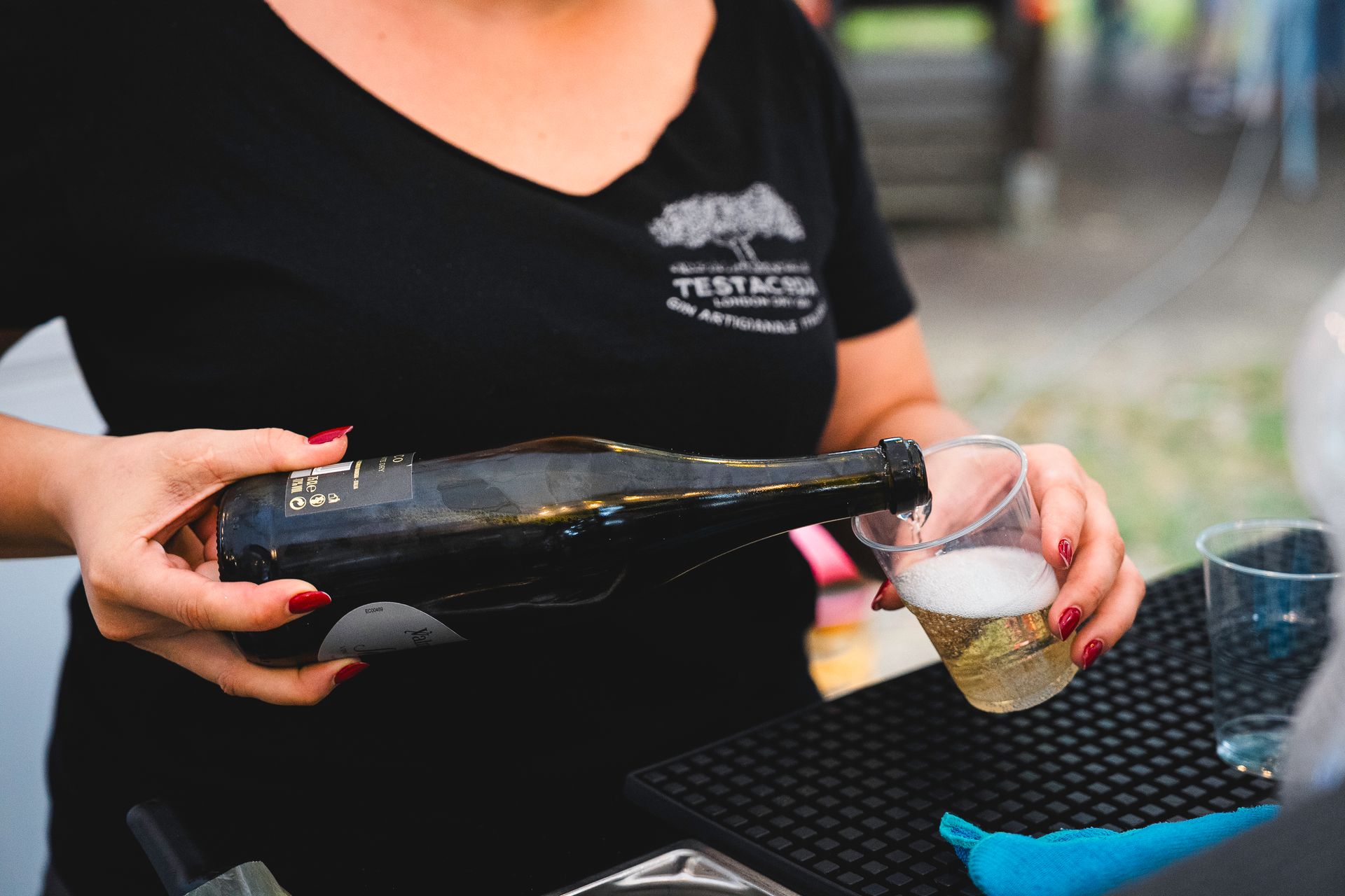 Persona con camicia nera che versa una bevanda da una bottiglia scura in un bicchiere di plastica trasparente.