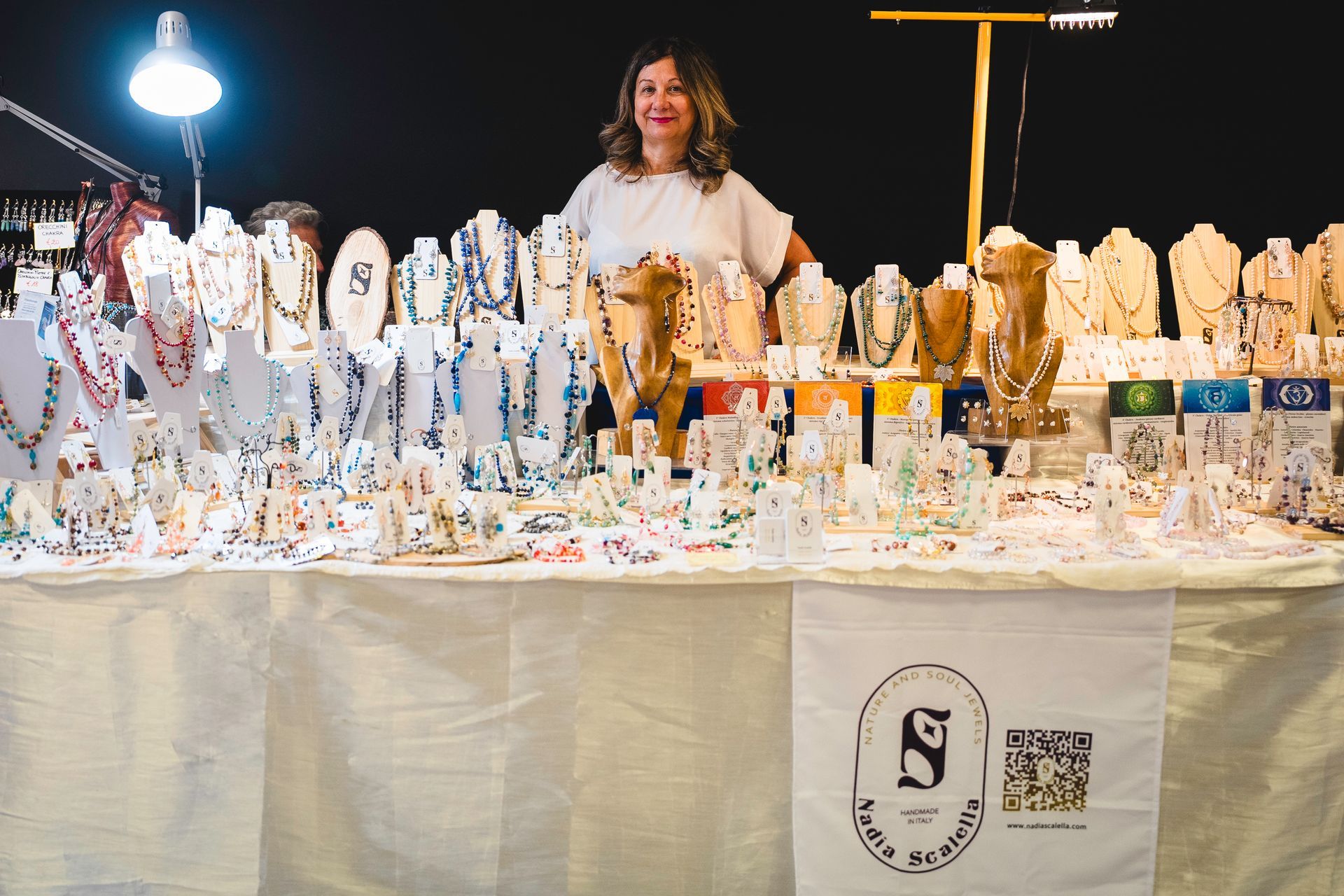 Donna sorridente alla bancarella di un mercato di gioielli. Molte collane e bracciali esposti.