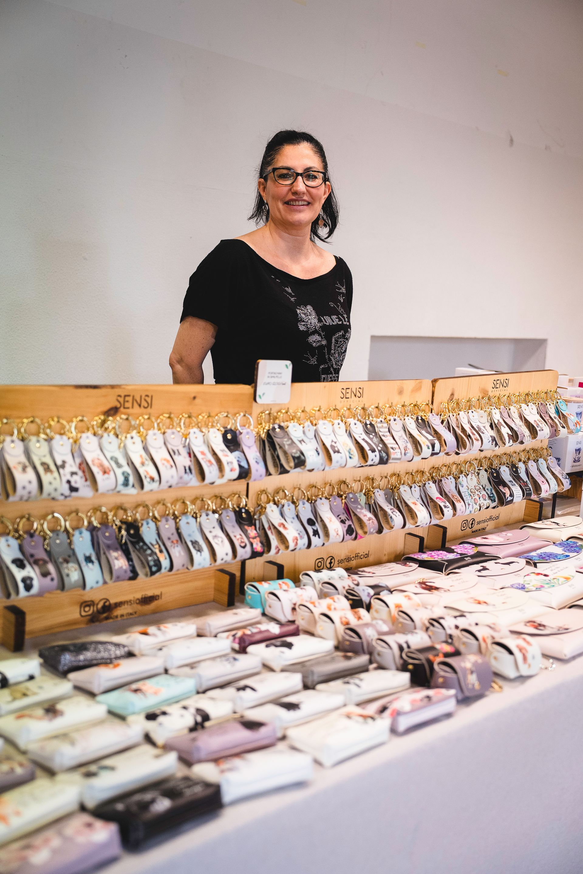 Una donna dietro un tavolo da lavoro espone vari accessori in tessuto; sorride alla telecamera.