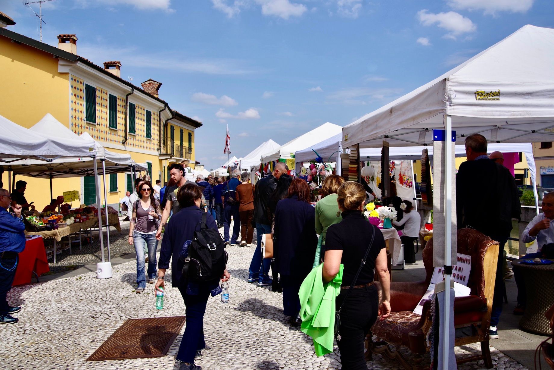 Mercato all'aperto con persone che curiosano tra le bancarelle sotto tende bianche su una strada acciottolata.