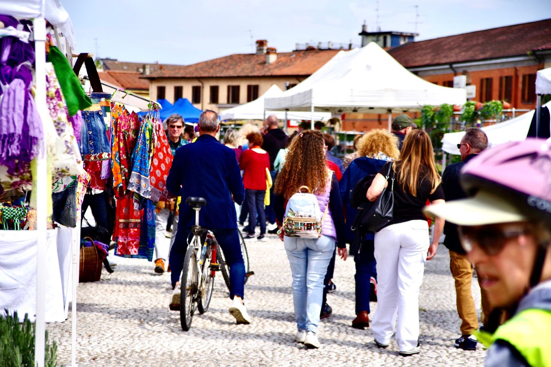 Mercato all'aperto con acquirenti, venditori e tende su una piazza pavimentata; edifici sullo sfondo.