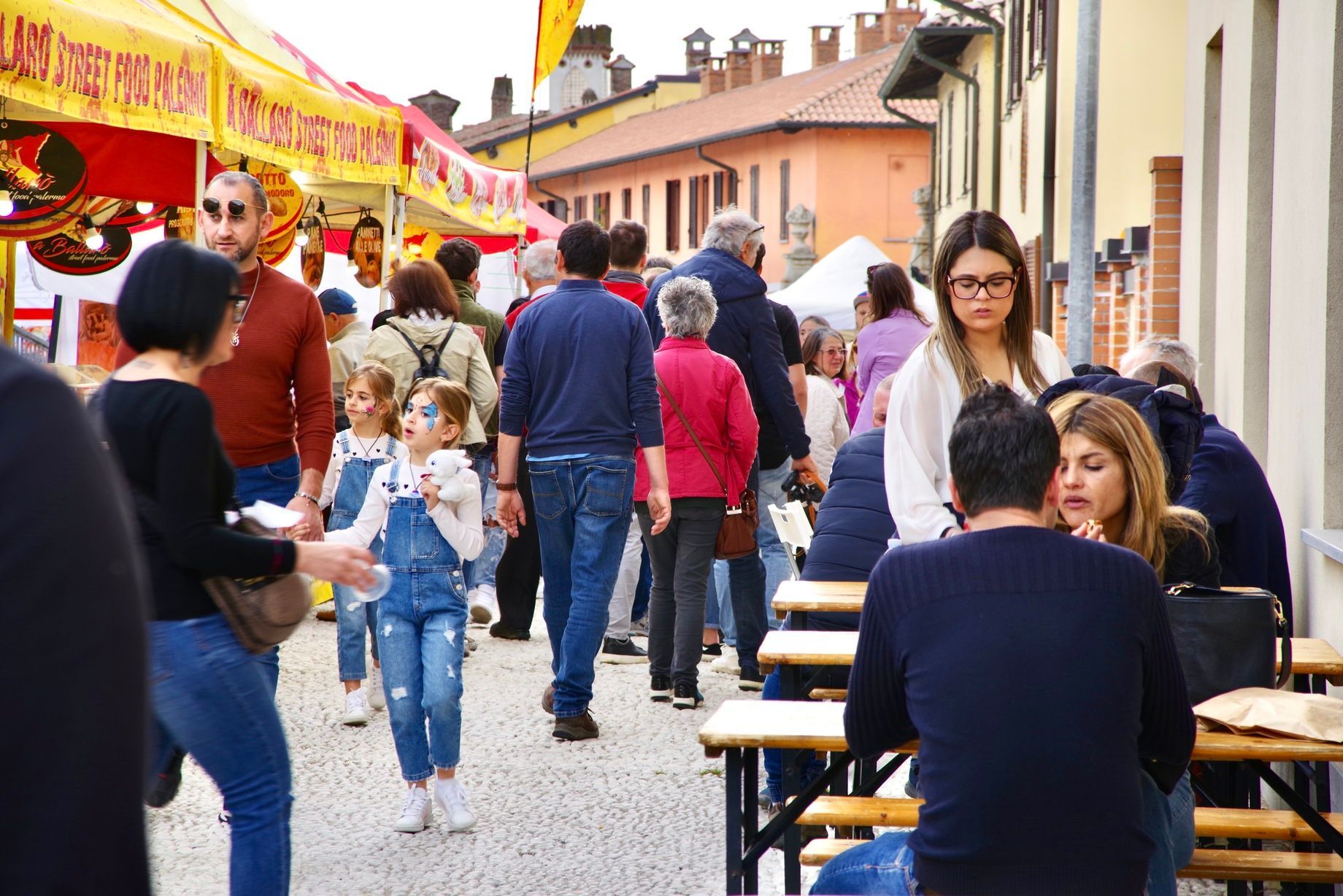 Folla a un festival di strada. Persone che camminano, chiacchierano e siedono ai tavoli. Bancarelle colorate fiancheggiano la strada.