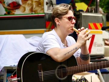 Donna con i capelli corti, che suona un flauto di legno ed è seduta con una chitarra acustica nera.