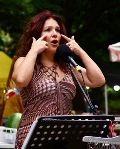 Una donna parla al microfono, toccandosi gli occhi. Indossa un abito a fantasia e si esibisce all'aperto.