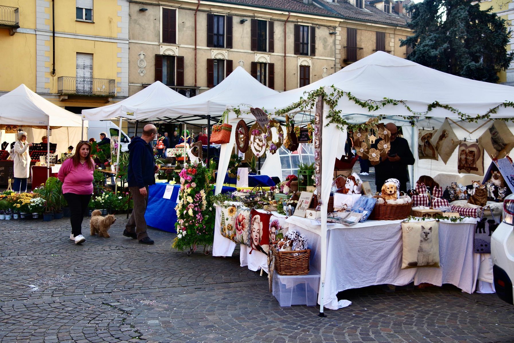 Mercato all'aperto con tende bianche, dove si vendono artigianato e fiori, in una piazza cittadina. Persone che curiosano.
